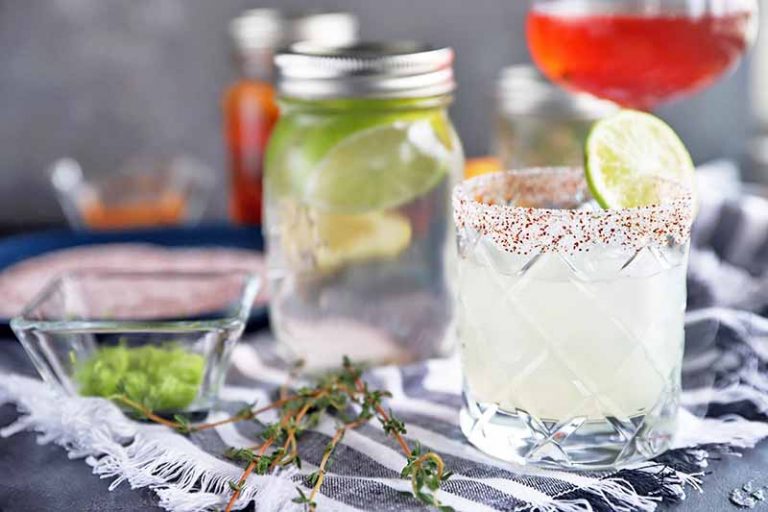 Horizontal image of a cocktail in a lowball glass in the foreground, garnished with a wedge of lime and a spiced salt rim, on a gray surface topped with a gray and white striped cloth with fringe,