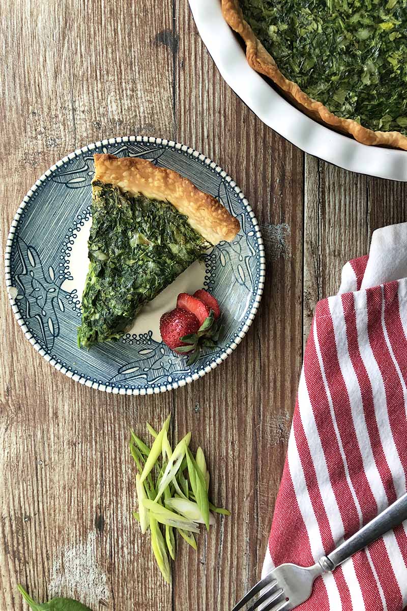 Vertical top-down image of a slice of a savory quiche with a green filing on a bowl with strawberries, next to a red and white towel, sliced scallions, and a white ceramic pan.