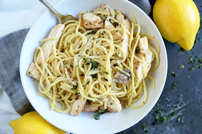 Overhead closely cropped horizontal image of a bowl of spaghetti with chicken, with two lemons on a white and gray cloth, on top of a dark gray surface.
