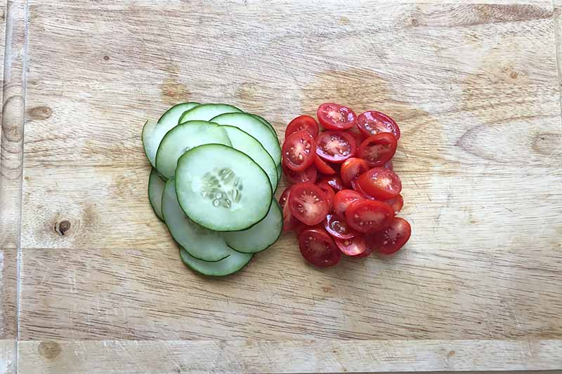 Horizontal image of sliced cucumbers and tomatoes on a wooden cutting board.