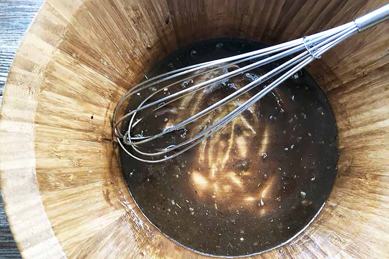 Horizontal image of a whisk resting in a brown liquid mixture in a wooden bowl.