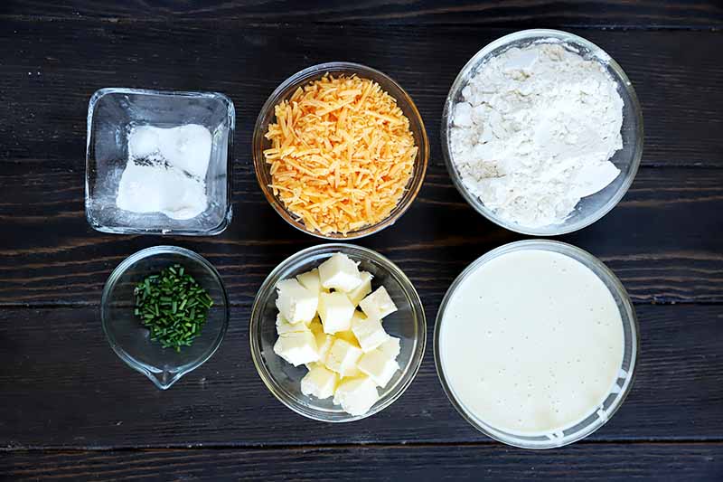 Horizontal image of bowls of dry ingredients, chives, shredded cheese, butter, and a sourdough starter on a black wooden surface.