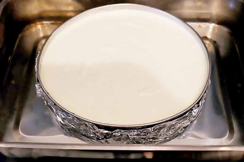 Horizontal image of a white unbaked dessert in a circular pan lined with aluminum foil in a water bath in the oven.