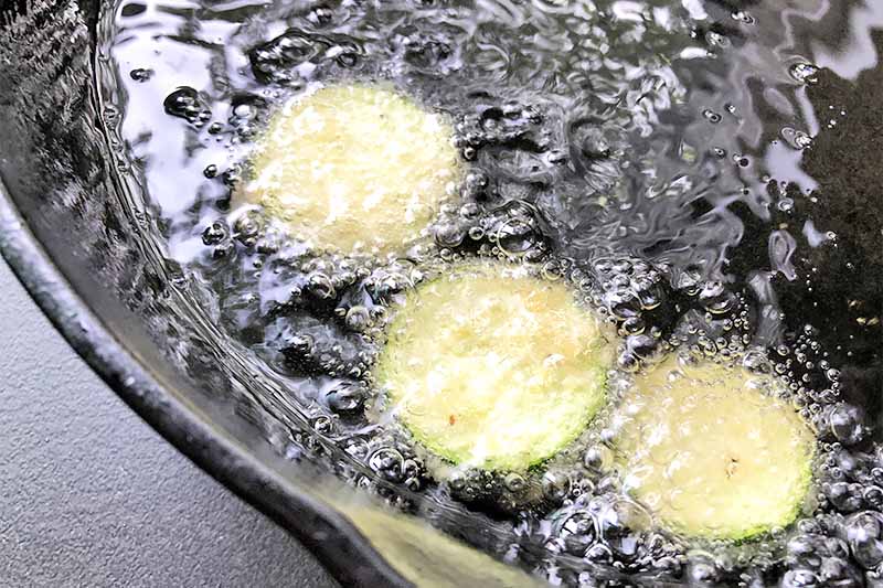 Horizontal image of bubbling oil and three slices of squash in a black pan.