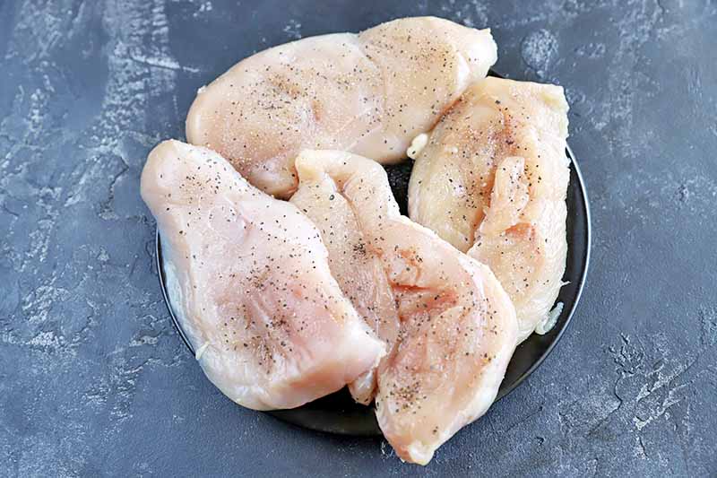 Horizontal overhead image of a small black plate filled with four boneless, skinless chicken breasts that have been sprinkled with salt and pepper, on a mottled gray background.