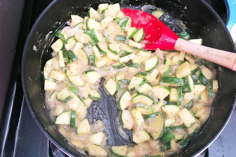 Horizontal image of cooking chopped zucchini and a creamy sauce in a pan with a red spatula.