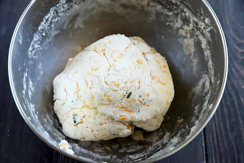 Horizontal image of a mass of dough speckled with cheddar cheese and herbs in a metal bowl.