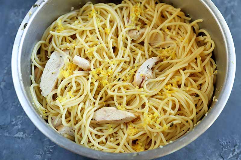 Horizontal overhead image of a metal bowl of spaghetti tossed with lemon sauce and grilled chicken, on a gray surface.
