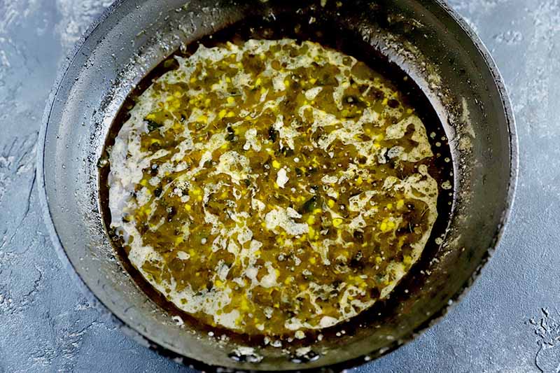 Horizontal overhead image of a saucepan filled with olive oil, garlic, herbs, and lemon juice, on a mottled gray surface.