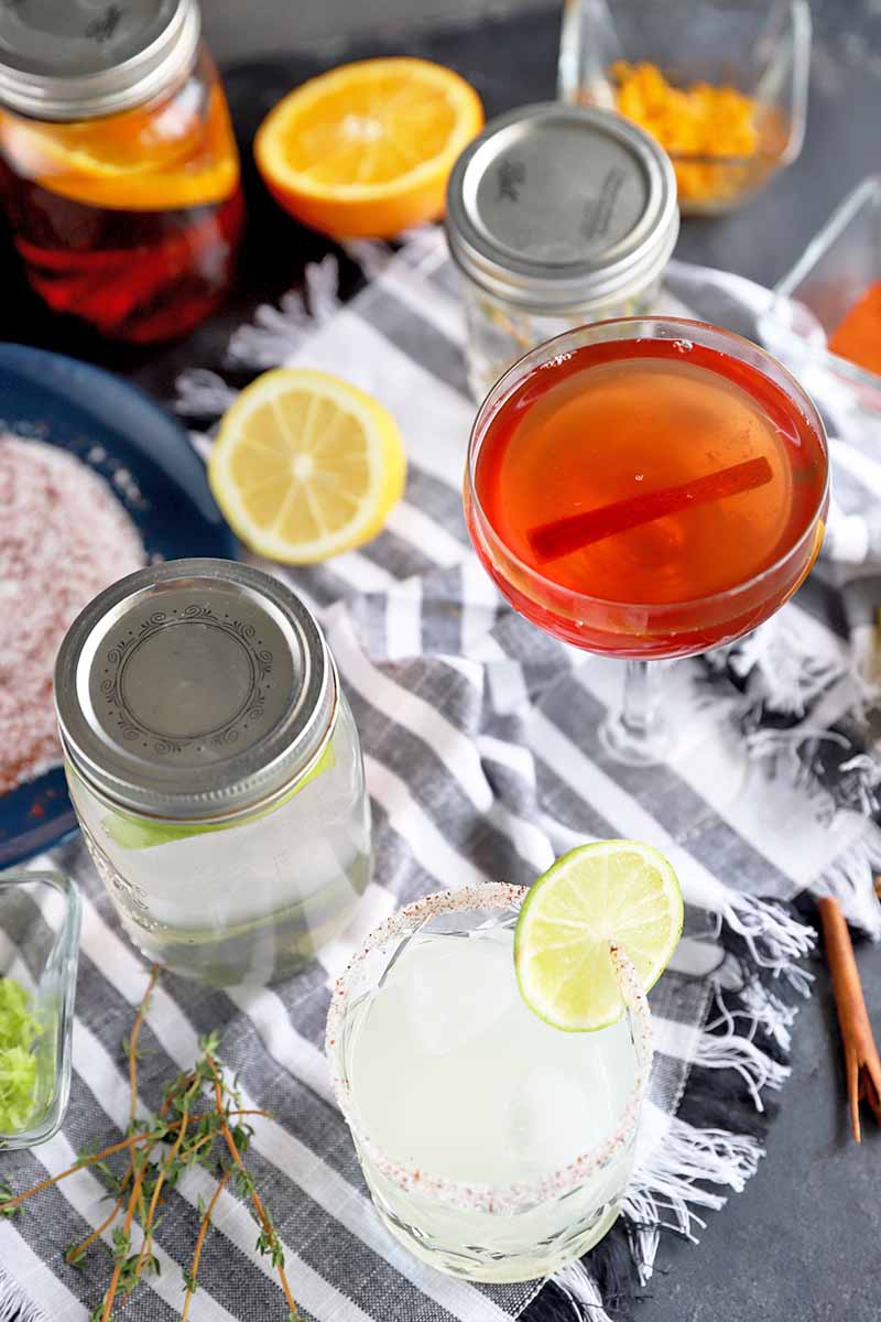 Vertical oblique overhead image of one bourbon and one tequila cocktail with garnishes, on a white and gray striped cloth topped with citrus, rimming salt, herbs, and whole spices, with jars of flavor-infused liquor, on a gray surface.