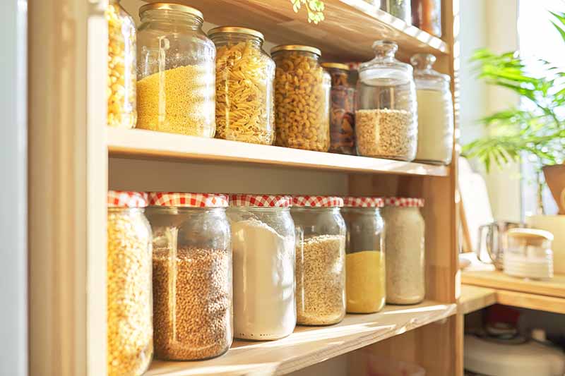 Horizontal image of shelves of yellow-themed dry goods in glass jars in a bright pantry.