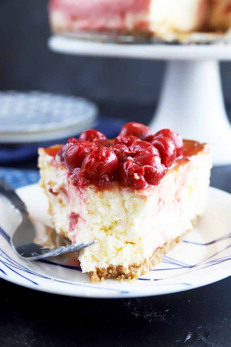 Vertical image of a cherry-topped cheesecake with a piece removed on a white plate next to a metal fork.