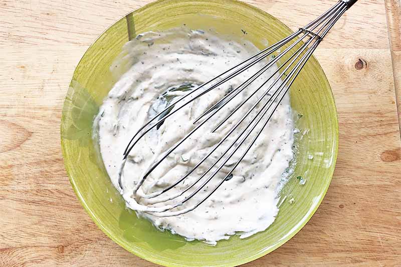 Horizontal image of a green bowl filled with a seasoned creamy mixture stirred by a metal whisk on a wooden surface.