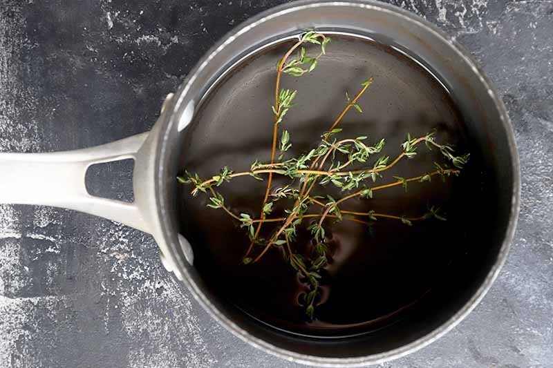 Horizontal overhead image of a small nonstick saucepan filled with simple syrup, with several sprigs of fresh thyme, on a gray surface streaked with white.