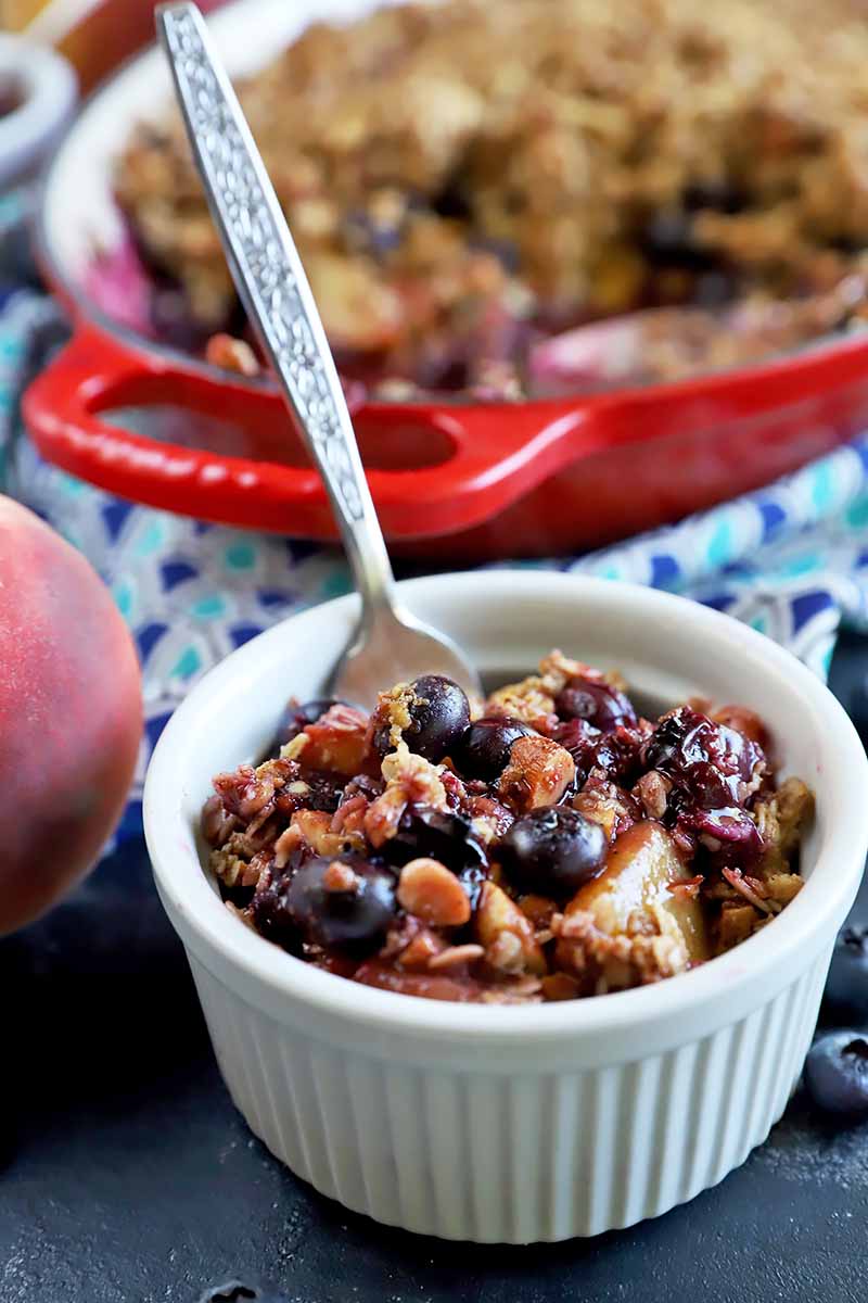 Vertical image of a white ramekin filled with a fruit and oat mixture with a spoon inserted into it, in front of a red oval pan with the same dessert on a bright blue towel with patterns.