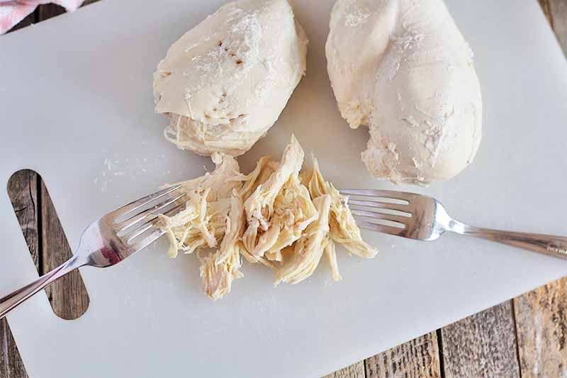 Horizontal image of shredded poached chicken breasts with two metal forks on a white cutting board that's on a wooden table.