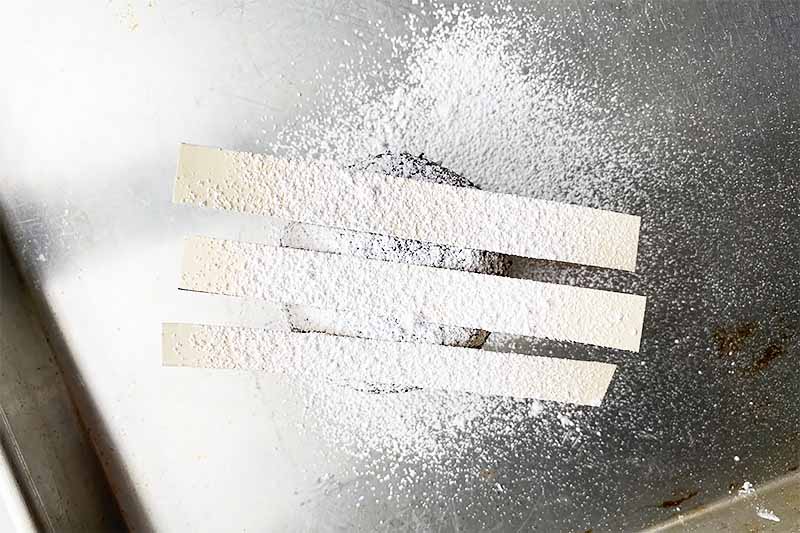 Horizontal image of a dusting of powdered sugar on three striped stencils on top of a dark circular dessert on a baking sheet.