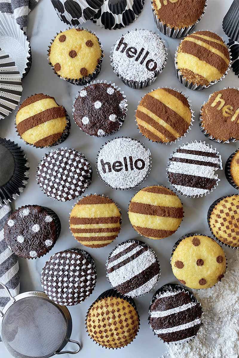 Vertical top-down image of assorted chocolate and vanilla cupcakes with decorations using powdered sugar and cocoa powder on a gray surface next to muffin liners and a mesh strainer.