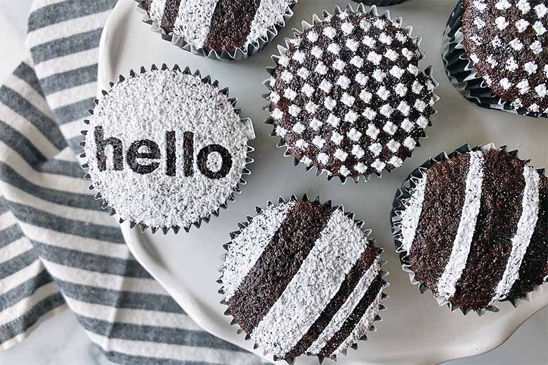 Horizontal top-down image of circular chocolate mini desserts decorated with powdered sugar in assorted patterns on a white stand on top of a striped black and white towel.