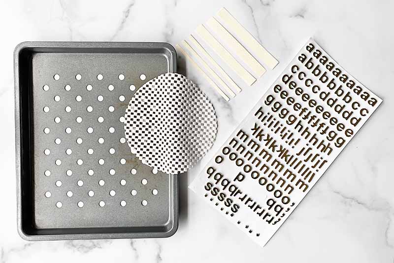 Horizontal image of a broiler pan, a jar grip, stencils, and a packet of alphabet stickers on a white marble surface.