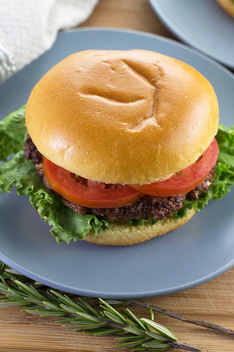 Vertical image of a neat sandwich with a fluffy bun, fresh tomato slices, a meat patty, and lettuce on a blue plate.