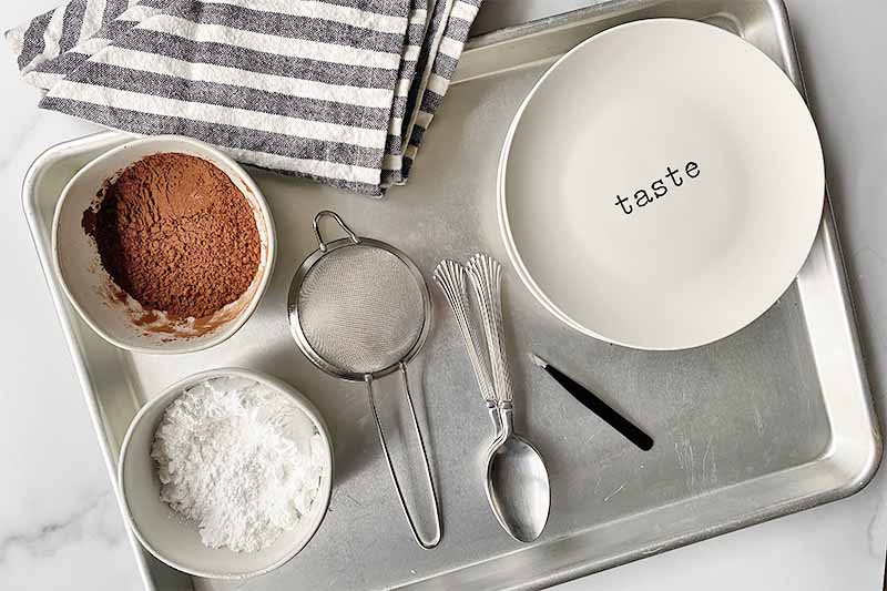 Horizontal image of bowls of powdered sugar and cocoa powder, a small mesh strainer, metal spoons, tweezer, and white plates on a baking sheet next to a black and white towel.