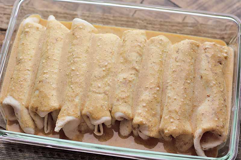 Horizontal image of a row of stuffed and rolled soft tortillas smothered in a light brown cream sauce in a glass casserole dish.