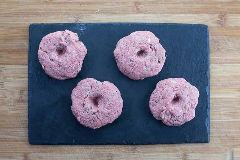 Horizontal image of four raw meat patties on a dark cutting board with imprints in the center of each one.