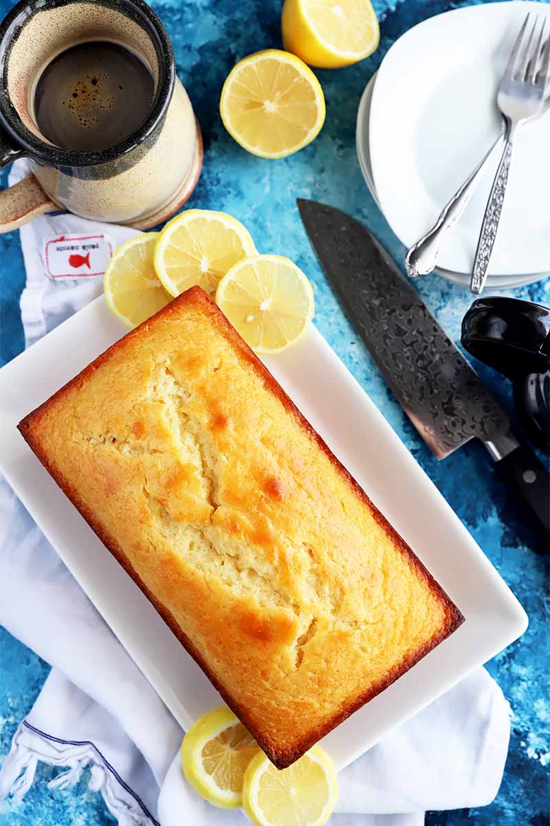 Vertical top-down image of a whole baked light yellow dessert in a loaf shape on a rectangular white plate surrounded by lemon slices, plates, silverware, a white towel, a mug, and a knife.