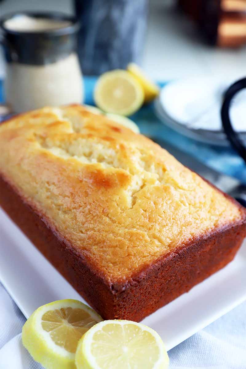 Vertical image of a whole baked light yellow loaf with browned edges on a white plate next to lemon wedges and a jar of sugar.