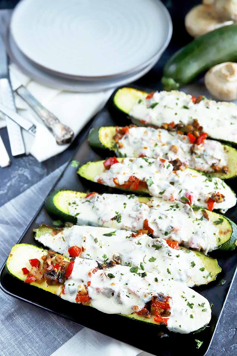 Vertical image of a black plate with a row of zucchini boats topped with melted cheese next to plates, silverware, and napkins.
