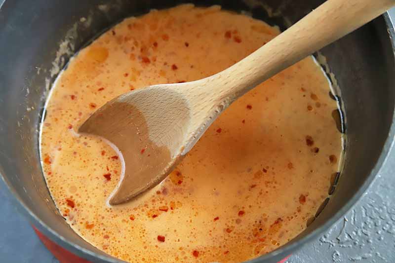 Horizontal image of a wooden spoon stirring a light red cream sauce in a black pot.