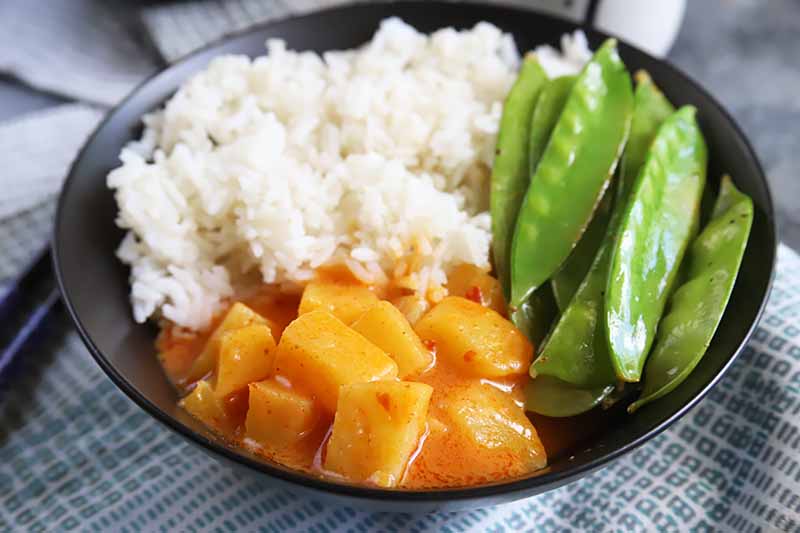 Horizontal image of a black bowl filled with rice, snap peas, and pineapple chunks in a creamy red sauce on a checkered blue towel.