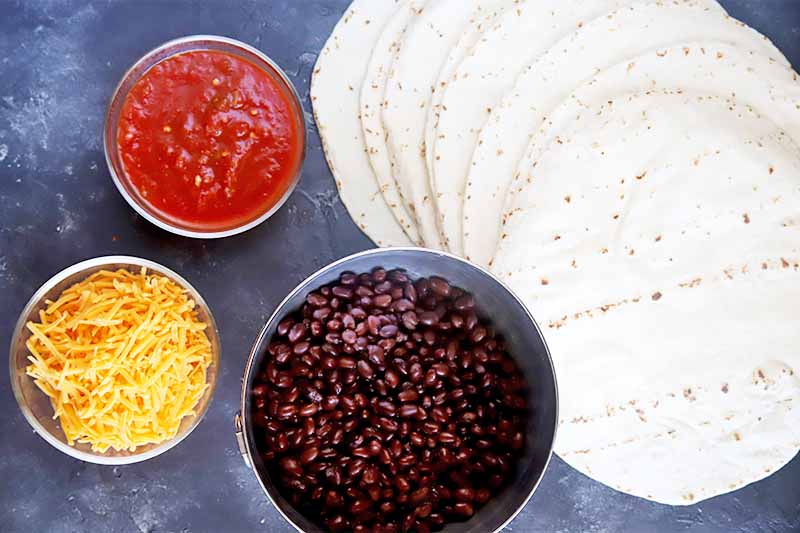 Horizontal image of a pile of flour tortillas and bowls of salsa, shredded cheddar cheese, and black beans on a dark surface.