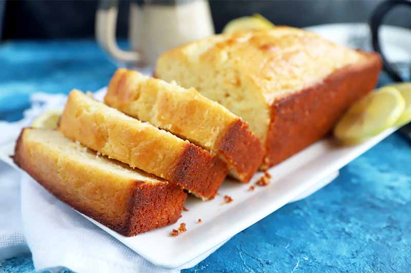 Horizontal image of a loaf baked good sliced into thick pieces on a white plate on a bright blue surface.