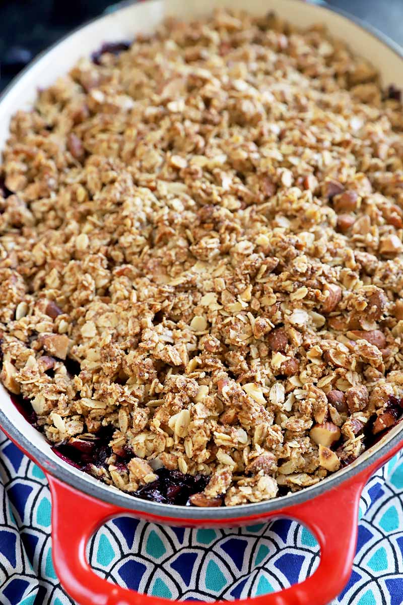 Vertical image of an oat and nut topping in an oval red pan on a towel with bright blue patterns.