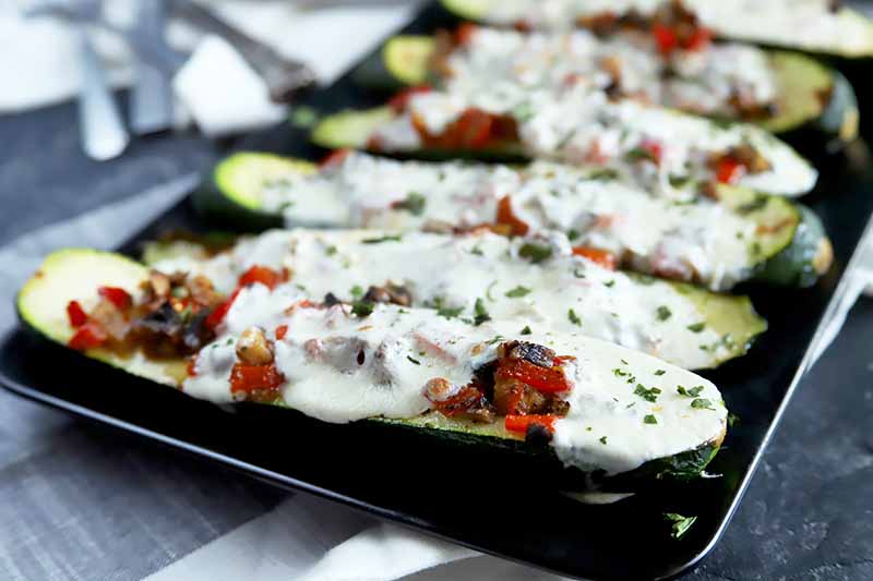 Horizontal image of a black plate with a row of zucchini boats topped with a vegetable and mozzarella mixture.