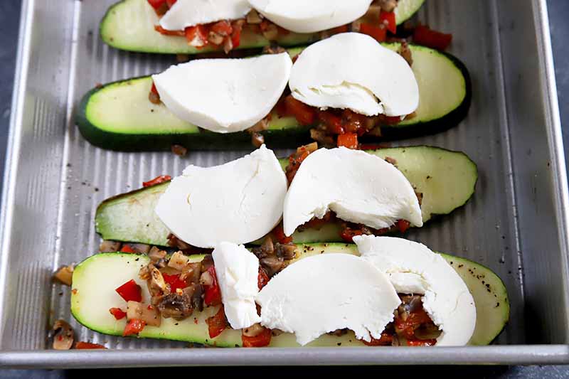 Horizontal image of slices of squash topped with a mushroom and pepper mixture and slices of mozzarella in a metal pan.