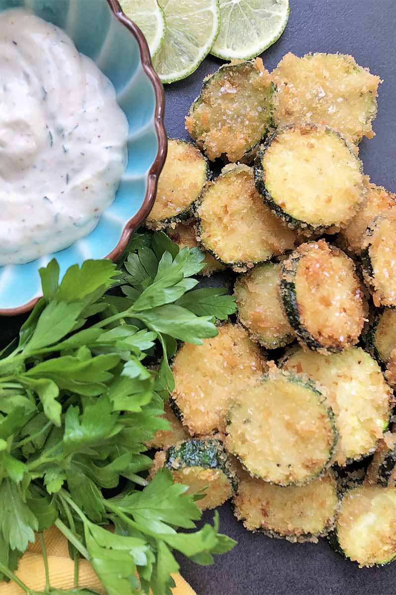 Vertical image of a pile of fried squash, fresh whole herbs, and a blue bowl filled with a creamy condiment on a dark plate.