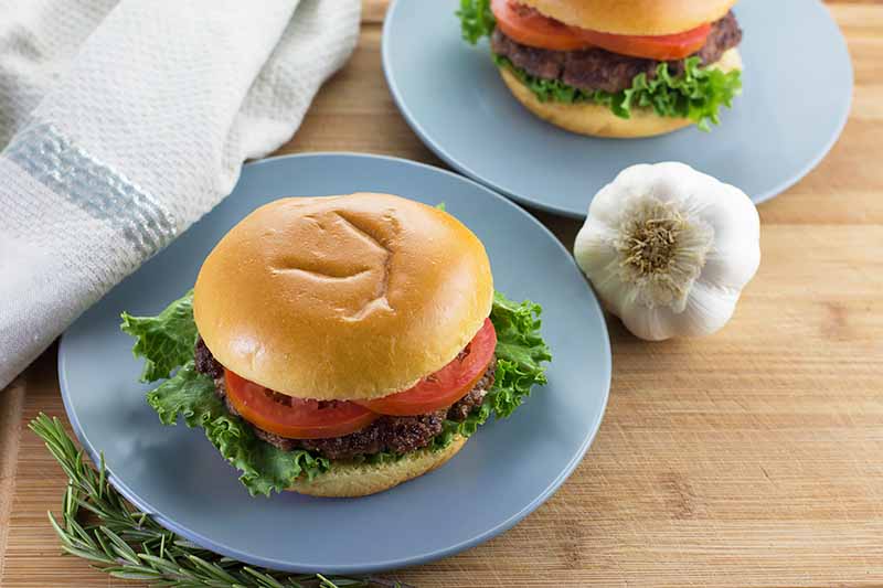 Horizontal image of two blue plates with burgers that have tomatoes, lettuce, and meat patties on a wooden surface next to a bulb of garlic.