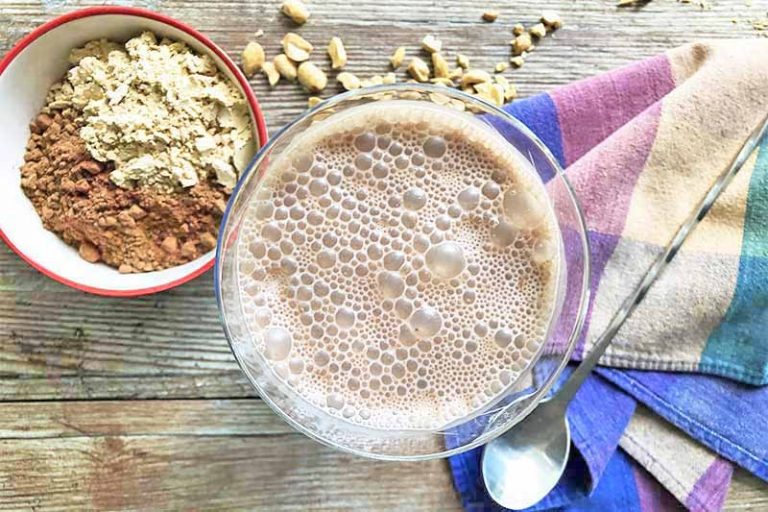 Horizontal image of a glass with a frothy light brown drink next to a colorful towel, a bowl of powdered ingredients, and a metal spoon.