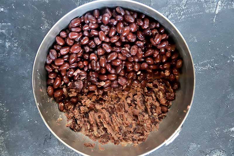 Horizontal image of a metal bowl with whole black beans, some partially crushed.