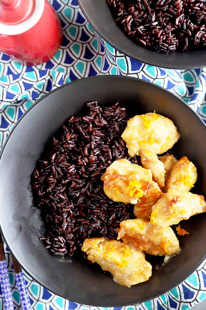 Vertical top-down image of a bowl of black rice with pieces of cooked chicken on the side, all on a patterned blue napkin.