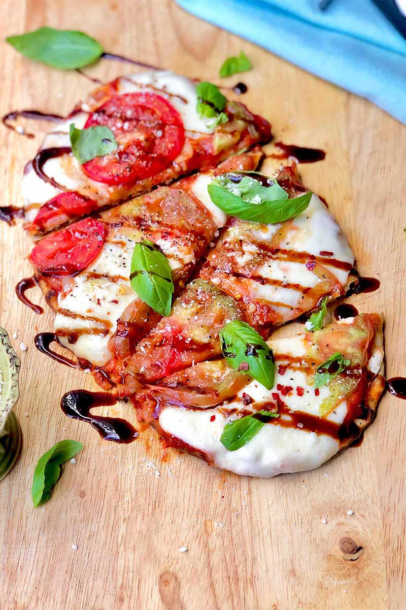 Vertical image of a sliced flatbread with cheese, tomato, and basil on a wooden cutting board.