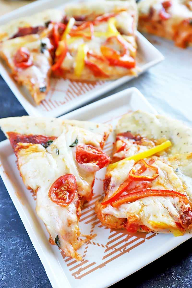 Vertical image of triangular slices of a baked herbed dough topped with cheese, tomato sauce, and whole tomatoes on square white plates.