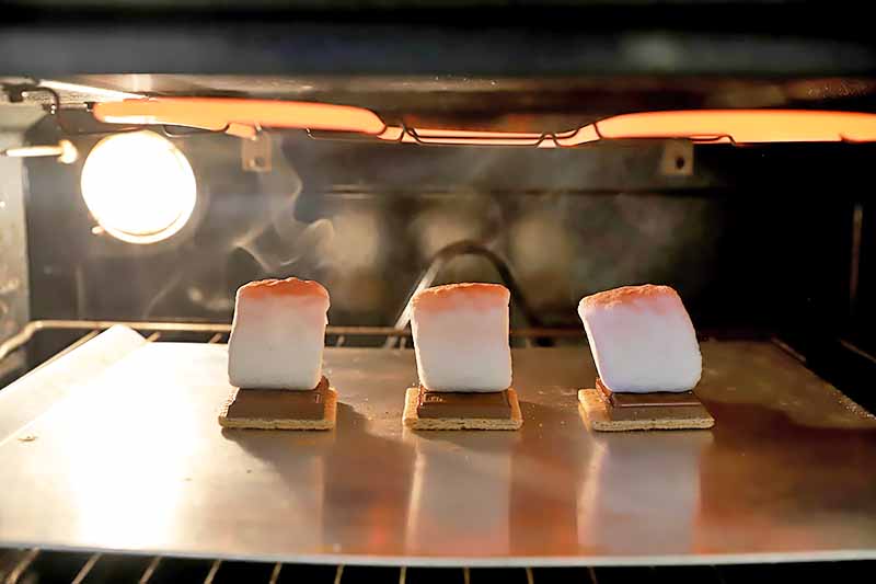 Horizontal image of broiling three graham crackers topped with chocolate and marshmallows that are browning on top.