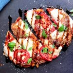 Horizontal top-down image of a Caprese flatbread on a dark surface next to a blue towel, basil, and marinara in a bowl.