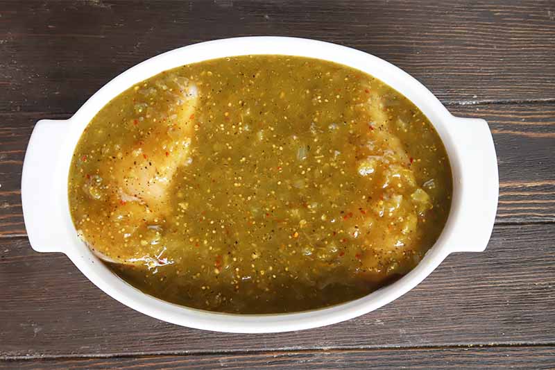 Horizontal image of a white casserole dish filled with meat covered in a green sauce on a dark surface.