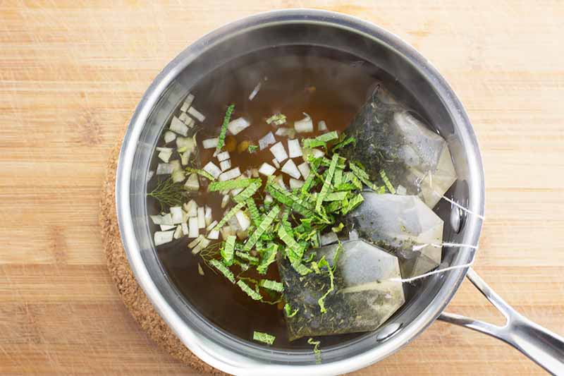 Horizontal image of a pot with tea bags and fresh chopped ingredients steeping in it on a wooden surface.