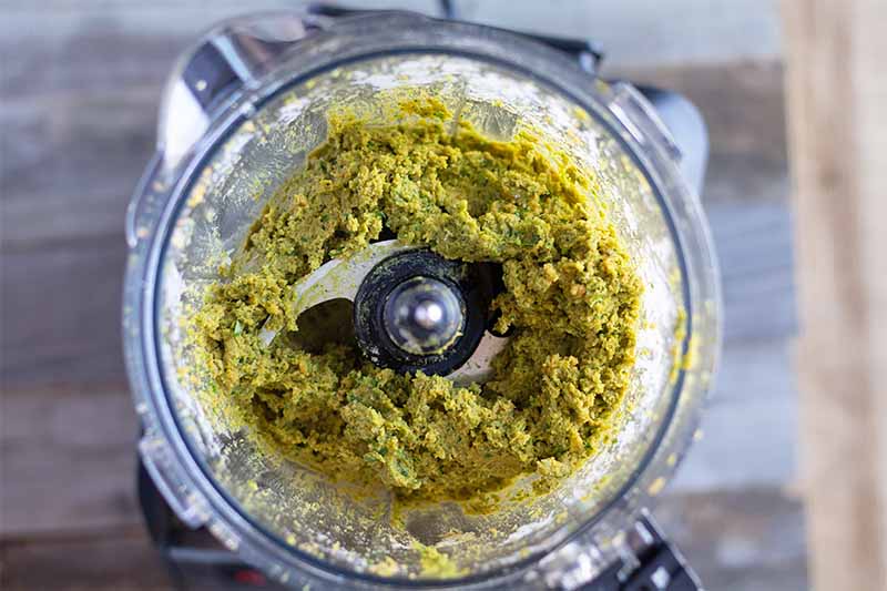 Horizontal image of a food processor with a mushy, grainy green mixture.